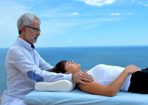 Javier de Maria en la playa haciendo Terapia Craneosacral con mujer en camilla en Alicante Explicacion de postura en camilla en el Curso de Terapia